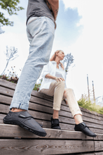 Load image into Gallery viewer, Two people wearing leguano neron shoes in sleek black, sitting on wooden steps in a natural setting.
